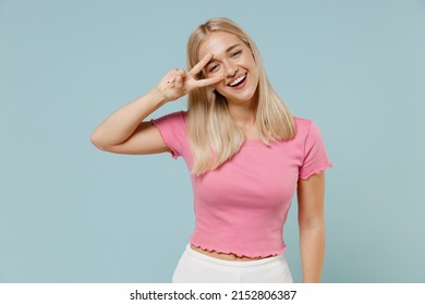Young caucasian smiling blonde woman 20s wear casual pink t-shirt showing victory sign cover eye with v-sign isolated on plain pastel light blue background studio portrait. People lifestyle concept