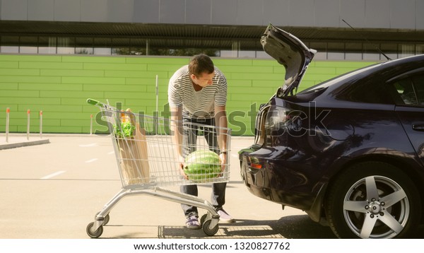 Young caucasian smiling male putting paper bags\
with food and watermelon into car trunk and sits in a car. Guy with\
foodstuffs near grocery\
store.