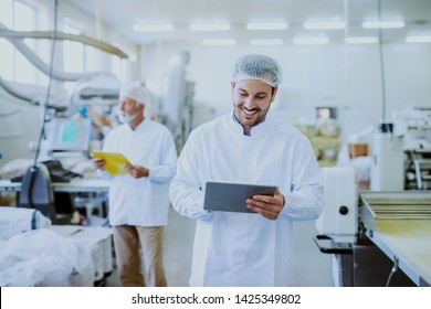 Young Caucasian Smiling Supervisor In Sterile White Uniform Using Tablet While Standing In Food Plant. In Background Older One Worker Controlling Machine.