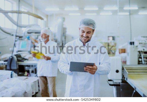 Young Caucasian smiling supervisor\
in sterile white uniform using tablet while standing in food plant.\
In background older one worker controlling\
machine.