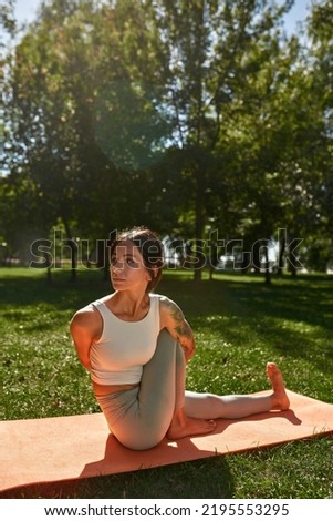 Young caucasian sportswoman practicing yoga in sage marichi's pose in sunny park. Girl with tattoos wearing sportswear sitting on fitness mat on green meadow. Concept of healthy lifestyle