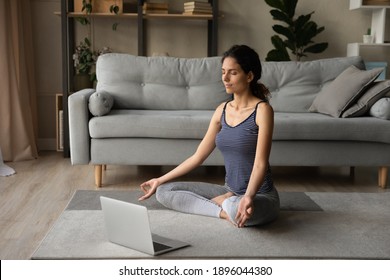 Young Caucasian sporty young woman sit on floor in living room practice yoga on online computer lesson. Millennial calm female meditate train at home watching having class on webcam on laptop.