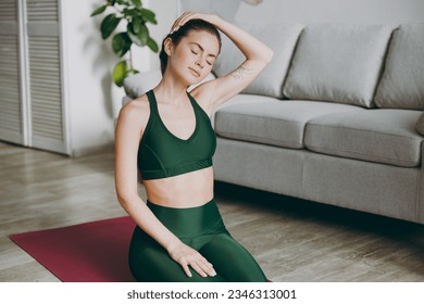 Young caucasian strong sporty athletic fitness trainer instructor woman wear green tracksuit holding head move it close eyes training do exercises at home gym indoor. Workout sport motivation concept