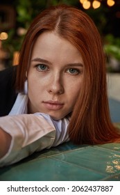 Young Caucasian Stylish Redhead Pretty Woman With Long Hair Lays Her Head On Her Arms And Table. Beautiful Pensive Melancholic Red Headed Lady Laying At Cafe. Lifestyle Closeup Portrait