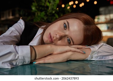 Young Caucasian Stylish Redhead Pretty Woman With Long Hair Lays Her Head On Her Arms And Table. Beautiful Pensive Melancholic Red Headed Lady Laying At Cafe. Lifestyle Portrait