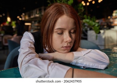 Young Caucasian Stylish Redhead Pretty Woman With Long Hair Lays Her Head On Her Arms And Table. Beautiful Pensive Melancholic Red Headed Lady Laying At Cafe With Closed Eyes. Lifestyle Portrait