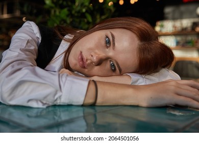 Young Caucasian Stylish Redhead Pretty Woman With Long Hair Lays Her Head On Her Arms And Table. Beautiful Pensive Melancholic Red Headed Lady Laying At Cafe. Lifestyle Portrait