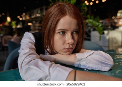 Young Caucasian Stylish Redhead Pretty Woman With Long Hair Lays Her Head On Her Arms And Table. Beautiful Pensive Melancholic Red Headed Lady Laying At Cafe. Lifestyle Portrait