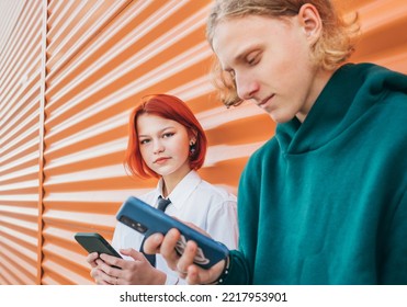 Young Caucasian Teen Girl Portrait Gazing At Camera. She Browsing Smartphone Devices With A Friend Boy Standing Near Orange Wall And. Careless Young Teenhood Time And A Modern Technology Concept Image