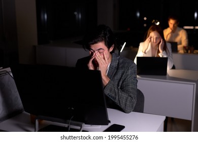 Young Caucasian Tired Brunnete Man In Suit Sits At Night In Office, Eyes Hurt From Overexertion, Suffering From Headache. Male Office Worker Does Not Meet The Deadline. Copy Space