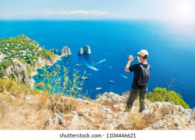 Young Caucasian Tourist Taking Picture Of Famous Faraglioni Rocks In Sea With His Smartphone On Top Of Mount Solaro After Hiking Up The Trail On Capri Island, Italy