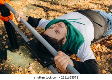 Young Caucasian Unshaven Man 34 Years Old Lies On Inclined Sloping Sports Board, Trains On Outdoor Sports Equipment On Sunny Autumn Day. Abs Workout, Leg Raises. Lifestyle Sport Portrait.