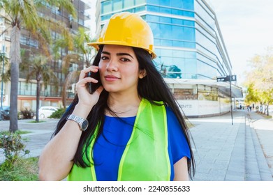 young caucasian venezuelan female engineer, wearing protective helmet and safety vest, standing outdoors talking on the phone and looking at the camera smiling, with a building behind her. - Shutterstock ID 2240855633