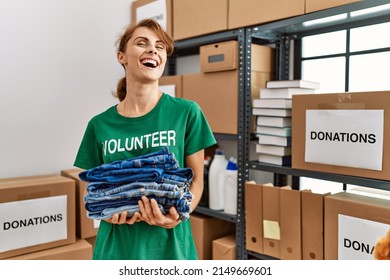 Young caucasian volunteer woman smiling happy holding stack of folded jeans at charity center.