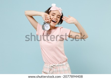 Young caucasian woman 20s wear pajamas jam sleep eye mask rest relaxing at home hold alarm clock stretch oneself in morning isolated on pastel blue background studio. Good mood night bedtime concept