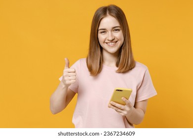 Young caucasian woman 20s wearing casual basic pastel pink t-shirt holding mobile cell phone surfing browsing internet showing thumb up like gesture isolated on yellow background studio portrait. - Shutterstock ID 2367234609
