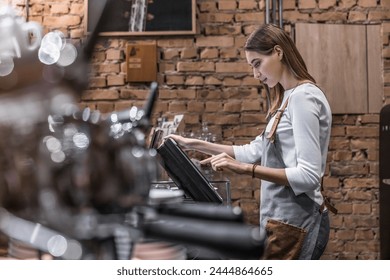 Young caucasian woman in apron working with cash box in coffee shop behind the counter. Female worker, barista, waitress, manager, administrator checking bill order on digital cash register. - Powered by Shutterstock