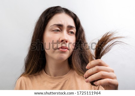 A young Caucasian woman in a beige t-shirt looks distressed at the dry ends of her long hair. White background
