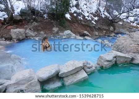 Young Caucasian woman in a black bikini stands in a hot spring pond and observes the snowy nature. Female traveler relaxes in the soothing hot water of Diamond Fork Hot Springs in the Utah wilderness.
