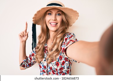 Young caucasian woman with blond hair wearing summer hat taking a selfie smiling with an idea or question pointing finger with happy face, number one 