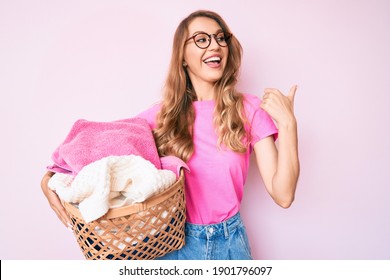 Young Caucasian Woman With Blond Hair Holding Laundry Basket Pointing Thumb Up To The Side Smiling Happy With Open Mouth 