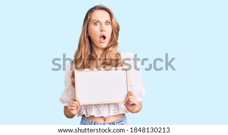 Young caucasian woman with blond hair holding blank empty banner scared and amazed with open mouth for surprise, disbelief face 
