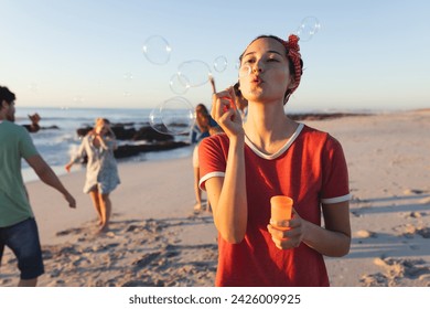 Young Caucasian woman blows bubbles on a beach at sunset. Friends enjoy a playful moment in the background, creating a relaxed outdoor atmosphere. - Powered by Shutterstock