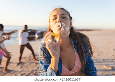 Young Caucasian woman blows bubbles on a sunny beach. Friends enjoy a playful moment in the background, highlighting leisure at the seaside. - Powered by Shutterstock