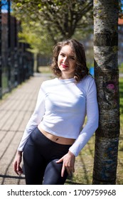 A Young Caucasian Woman With Brown Hair And Green Eyes Is Posing By The Blooming Flower Tree By The Football Field In The Spring And Wearing White Long Sleeved Shirt, Black Tight Leggings, Boots 