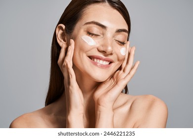 A young Caucasian woman with brunette hair smiles brightly while delicately touching her face with cream - Powered by Shutterstock