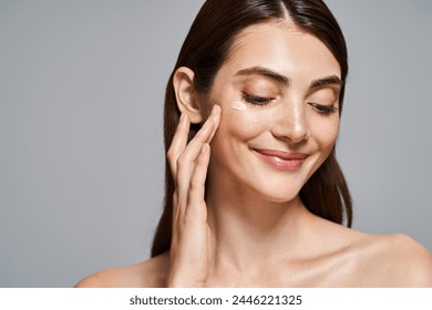 A young Caucasian woman with brunette hair smiling and gently touching her face with cream - Powered by Shutterstock