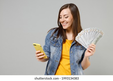 Young caucasian woman in casual trendy denim jacket yellow t-shirt hold mobile cell phone fan of cash money in dollar banknotes isolated on grey background studio portrait. People lifestyle concept