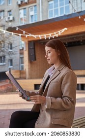 Young Caucasian Woman In Coat Using Tablet Outdoors On Sunny Day, Reading Ebook, Watching Movie. Attractive Female Street Portrait At Fall Or Spring. Autumn Lifestyle, Modern Communication Technology