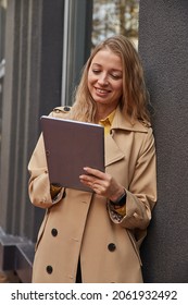 Young Caucasian Woman In Coat Using Tablet Outdoors On Sunny Day, Reading Ebook, Watching Movie. Attractive Female Street Portrait At Fall Or Spring. Autumn Lifestyle, Modern Communication Technology