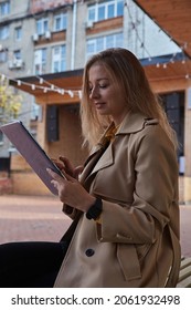 Young Caucasian Woman In Coat Using Tablet Outdoors On Sunny Day, Reading Ebook, Watching Movie. Attractive Female Street Portrait At Fall Or Spring. Autumn Lifestyle, Modern Communication Technology