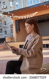 Young Caucasian Woman In Coat Using Tablet Outdoors On Sunny Day, Reading Ebook, Watching Movie. Attractive Female Street Portrait At Fall Or Spring. Autumn Lifestyle, Modern Communication Technology