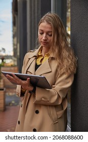 Young Caucasian Woman In Coat Using Tablet Outdoors On Sunny Day, Reading Ebook, Watching Movie. Attractive Female Street Portrait At Fall Or Spring. Autumn Lifestyle, Modern Communication Technology