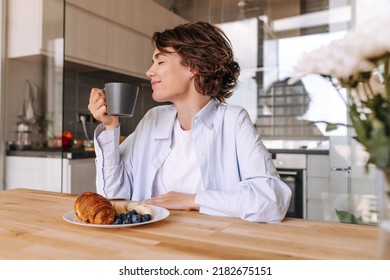 Young Caucasian Woman With Cup Coffee In Home. In Hands Black Cup Of Coffee Or Tea, Smiling With Close Eyes Sitting At The Kitchen. Consept Of Enjoying Life