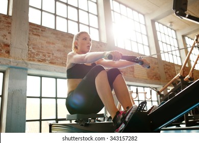 Young Caucasian Woman Doing Exercises On Fitness Machine In Gym. Female Using Rowing Machine At  Fitness Club.