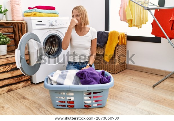 Young caucasian woman\
doing laundry with clothes in the basket smelling something stinky\
and disgusting, intolerable smell, holding breath with fingers on\
nose. bad smell 