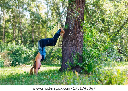Young caucasian woman doing yoga exercises in summer city park. Woman doing handstand next to the tree