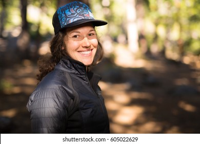 Young caucasian woman dressed in outdoors clothing stands in a forest and smiles at viewer