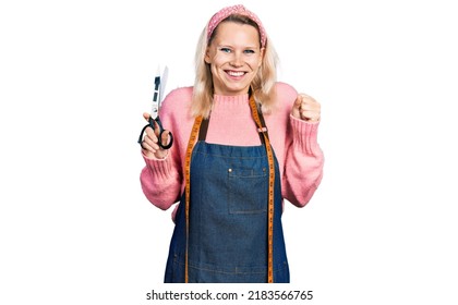 Young Caucasian Woman Dressmaker Designer Wearing Atelier Apron Holding Scissors Screaming Proud, Celebrating Victory And Success Very Excited With Raised Arm 