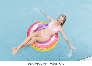 Young Caucasian woman enjoys a sunny day floating in a pool on a colorful ring. She wears a pink polka-dot bikini, her blonde hair tied up, conveying a relaxed vacation vibe. - Powered by Shutterstock