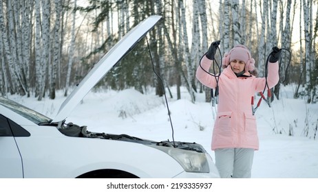 Young Caucasian Woman Examining Booster Jumper Cables While Standing By The Open Hood Of Her Car In Cold Winter Day