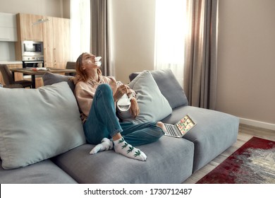Young caucasian woman exhaling the smoke while smoking marijuana from a bong or glass water pipe, sitting on the couch. Cannabis and weed legalization concept. Horizontal shot