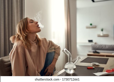 Young Caucasian Woman Exhaling The Smoke While Smoking Marijuana From A Bong Or Glass Water Pipe, Sitting In The Kitchen. Red Weed Grinder And Lighter On The Table. Cannabis Legalization Concept