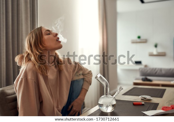 Young caucasian woman exhaling the smoke while\
smoking marijuana from a bong or glass water pipe, sitting in the\
kitchen. Red weed grinder and lighter on the table. Cannabis\
legalization concept