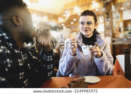 Young caucasian woman with eyeglasses drinking an hot chocolate at cafe with her African boyfriend - biracial couple at coffee shop having fun together - diverse ethnicity life style concept 