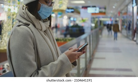 Young Caucasian Woman in Fashionable Clothes and Surgical Hygiene Mask Waiting in Shopping Mall Swiping Her Smartphone Looking for Best Offers on Christmas Season Sale Day